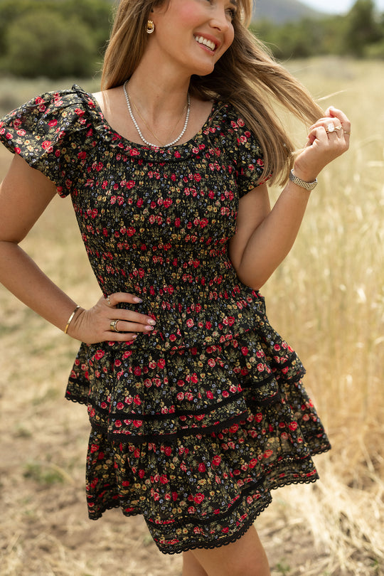 A woman stands in a grassy field, smiling in the Ivy City Co Annalise Top in Obsidian Vintage Poppies—a black top with a red floral pattern, layered skirt effect, short ruffled sleeves, and smocked bodice. One hand is on her hip, the other in her hair.
