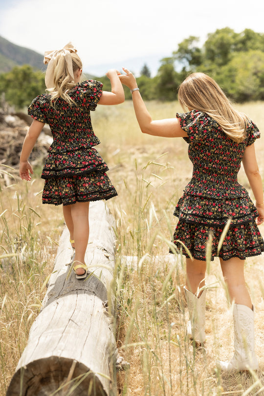 Two girls wearing matching Ivy City Co Annalise Tops in Obsidian Vintage Poppies, one in white cowboy boots with a hair bow and smocked bodice details, hold hands as they balance on a log in a sunny grassy field.