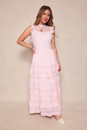 A woman poses against a beige background in the Annie Dress in Light Pink by Ivy City Co, featuring lace, ruffled layers, and a romantic silhouette. She finishes the look with tan pointed flats, gold jewelry, and long wavy light brown hair.