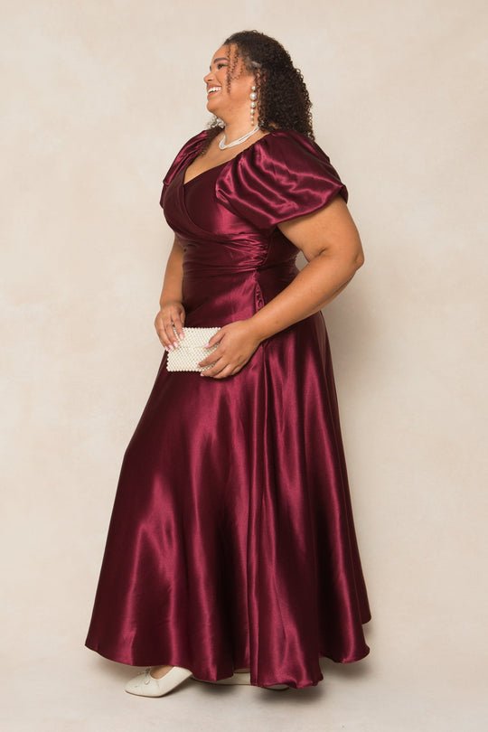 A woman stands smiling in the Ivy City Co Tessie Dress in Wine Satin, featuring puff sleeves. She accessorizes with a white clutch, white shoes, pearl earrings, and a necklace, posing against a neutral background.