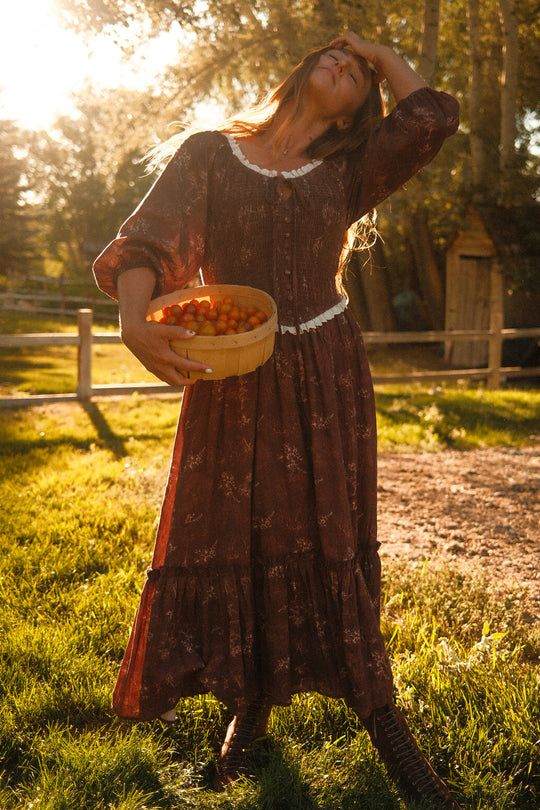 Basked in sunlight in a grassy yard with trees and a wooden fence, a woman stands wearing the Ivy City Co Asha Dress in Warm Cocoa Valencia Toile—a long, flowy cotton dress offering both style and comfort that’s even maternity friendly. She tilts her head back, touching her forehead while holding a basket of yellow-orange tomatoes.