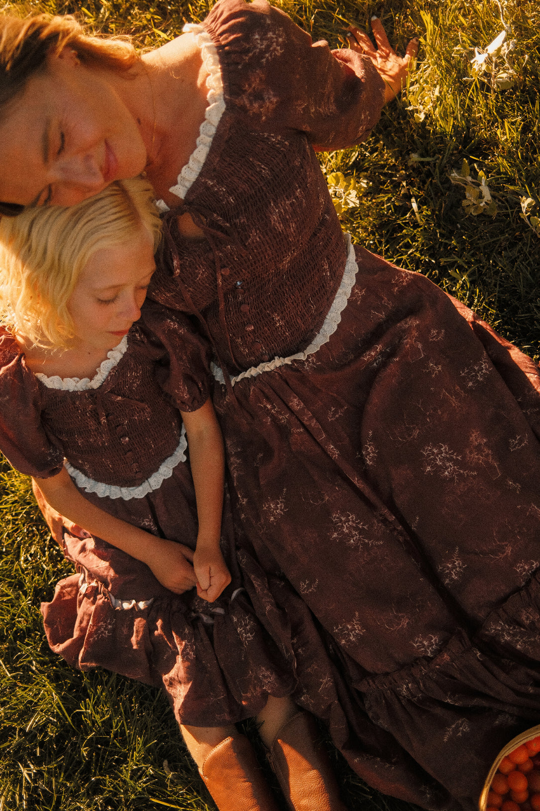 A woman and a child in matching Asha Dresses in Warm Cocoa Valencia Toile by Ivy City Co, featuring delicate white trim, sit on the grass together with eyes closed, soaking in a tranquil moment under the sun. Made from soft cotton, these maternity-friendly dresses provide comfort and style through every stage of motherhood.
