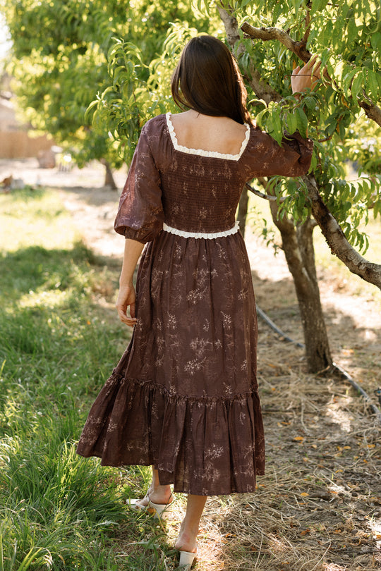 A woman stands outdoors by a tree, facing away and touching a branch, wearing the Ivy City Co Asha Dress in Warm Cocoa Valencia Toile—a vintage-inspired, long-sleeved cotton dress with white lace trim that features a graceful silhouette and is maternity friendly.