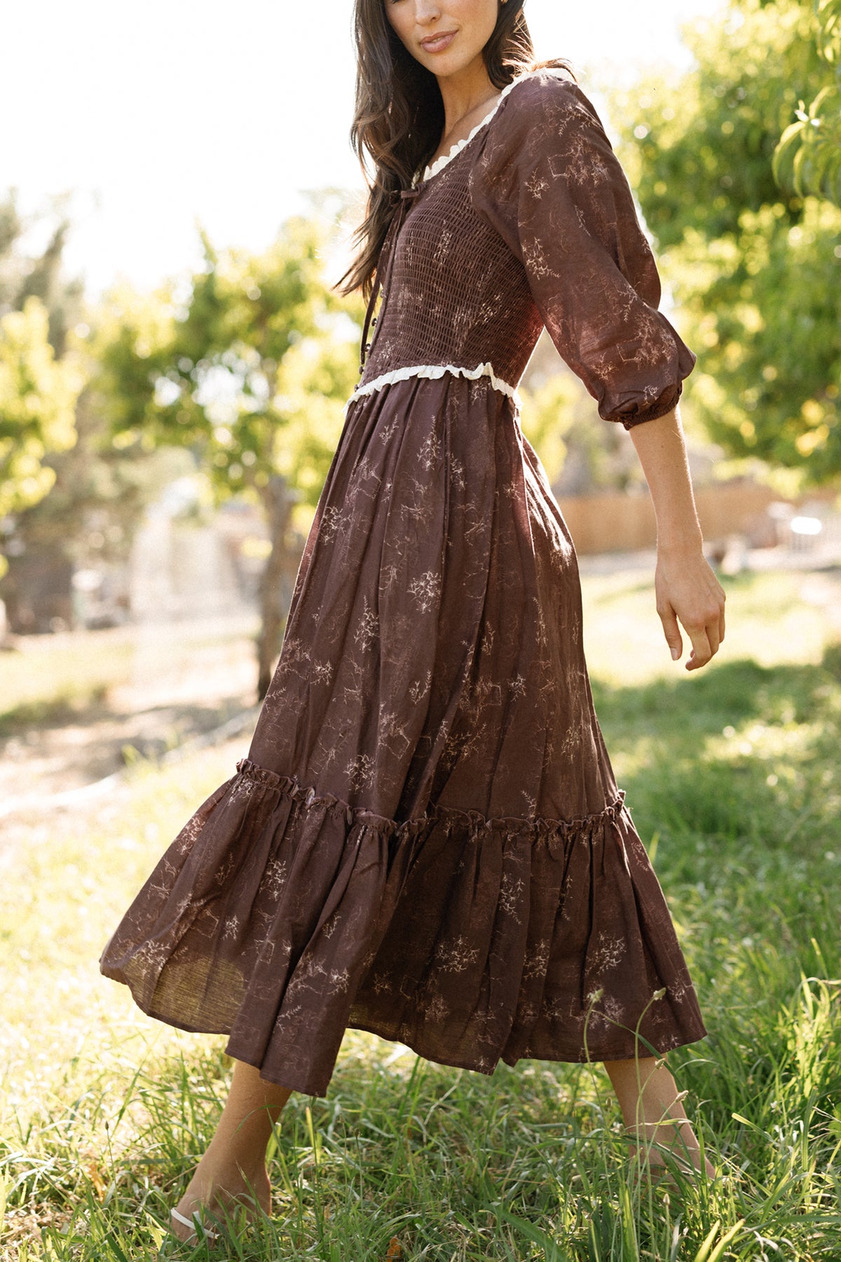 A woman walks barefoot on grass in a sunlit outdoor setting with trees in the background, wearing Ivy City Co’s Asha Dress in Warm Cocoa Valencia Toile—a long, brown floral cotton dress featuring puffed sleeves. This maternity-friendly style provides comfort and effortless elegance for warm, leisurely days.