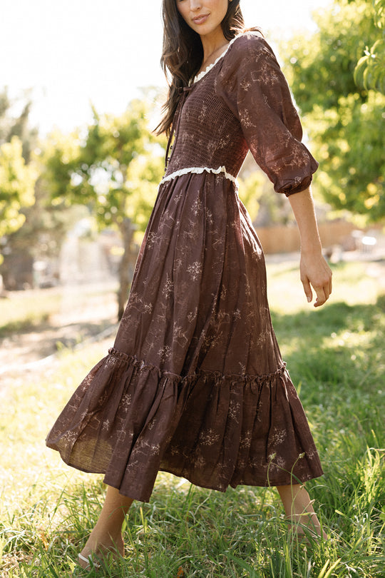 A woman walks barefoot on grass in a sunlit outdoor setting with trees in the background, wearing Ivy City Co’s Asha Dress in Warm Cocoa Valencia Toile—a long, brown floral cotton dress featuring puffed sleeves. This maternity-friendly style provides comfort and effortless elegance for warm, leisurely days.