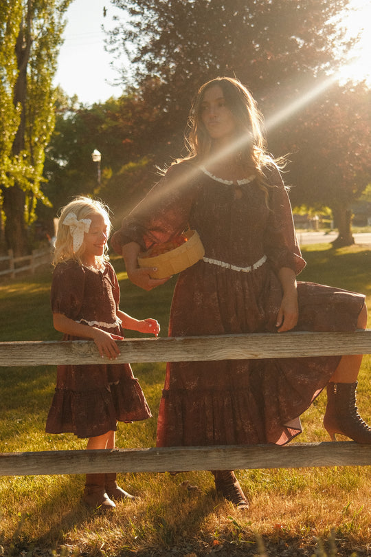 A woman and a girl stand by a wooden fence in sunlight, both dressed in the Ivy City Co Asha Dress in Warm Cocoa Valencia Toile—an effortlessly charming, maternity-friendly style perfect for idyllic days outdoors. The woman carries a basket, and trees bathed in golden sunlight fill the rustic background, completing the warm scene.