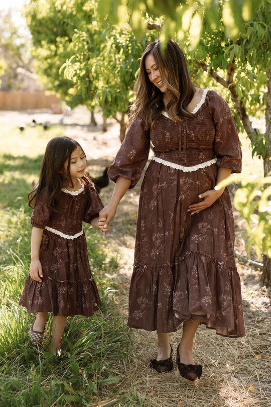 A woman and a young girl, both dressed in matching Ivy City Co Asha Dresses in Warm Cocoa Valencia Toile, hold hands and walk outdoors beneath green trees on a sunny day. These stylish, comfortable dresses are maternity-friendly—perfect for creating special moments together.