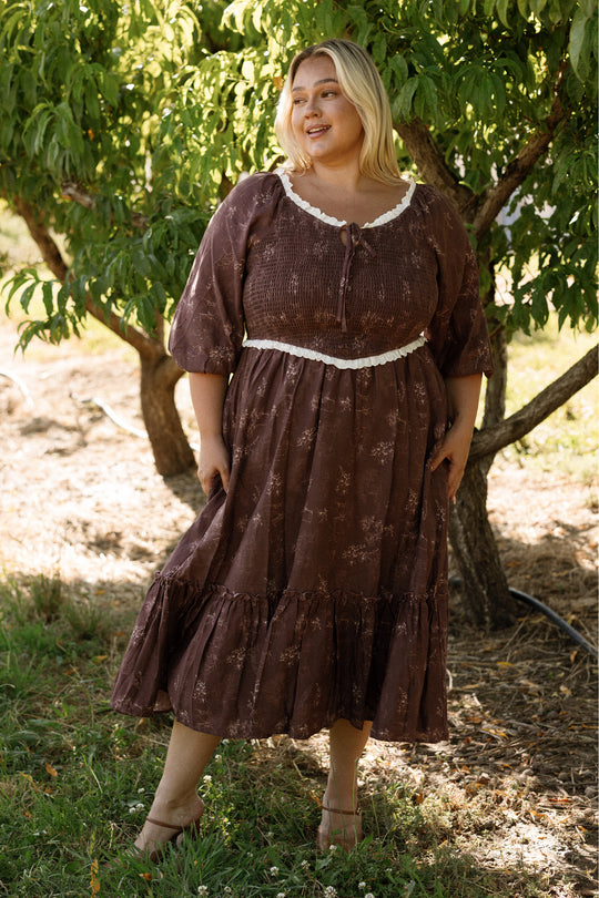 A smiling woman stands outdoors on a grassy lawn in front of leafy trees, enjoying a sunny day in the Asha Dress in Warm Cocoa Valencia Toile by Ivy City Co. The long-sleeved cotton dress features charming ruffles and is both stylish and maternity friendly, offering comfort and ease.