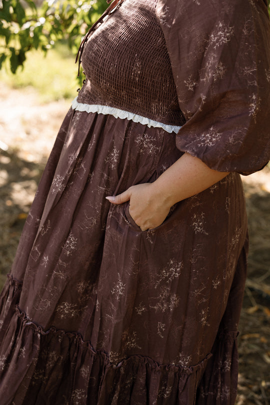 A person wearing the Ivy City Co Asha Dress in Warm Cocoa Valencia Toile, a brown floral-patterned cotton dress with puffed sleeves and a lace trim, is standing outdoors with one hand in the dress pocket. This style is also maternity friendly.