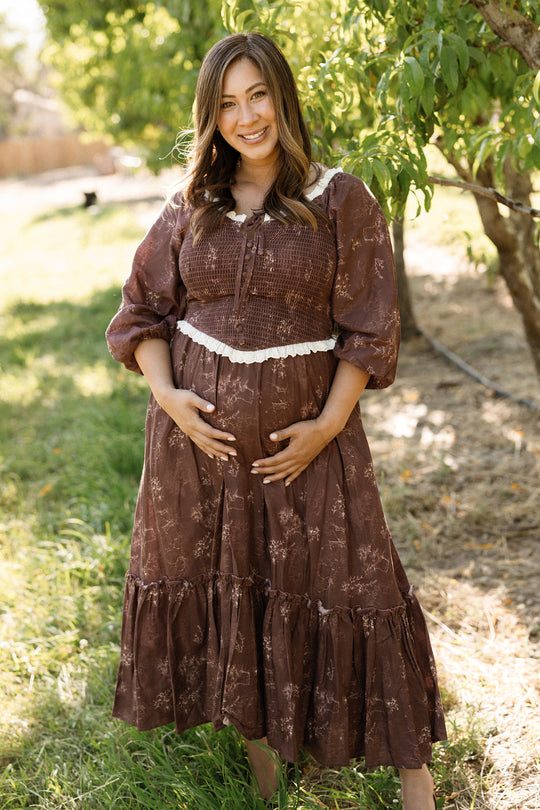 A pregnant woman wearing the Ivy City Co Asha Dress in Warm Cocoa Valencia Toile, a maternity-friendly cotton dress, stands outdoors on grass, smiling at the camera with her hands on her belly. Green trees and sunlight fill the background.