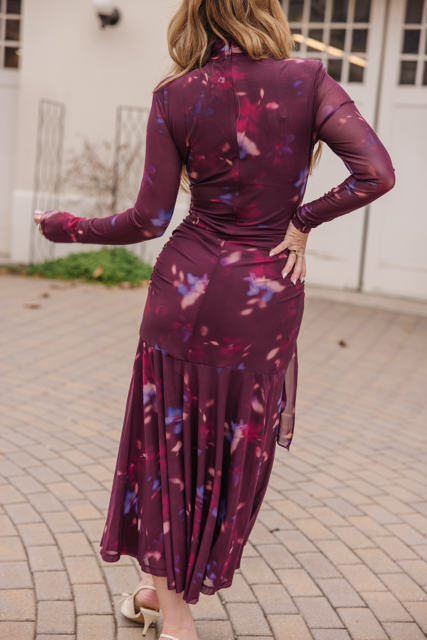 A woman stands on a paved surface, facing away from the camera, wearing the Ivy City Co Aubriella Dress in Burgundy Floral with a fitted bodice and pleated skirt, paired with light-colored heeled sandals.