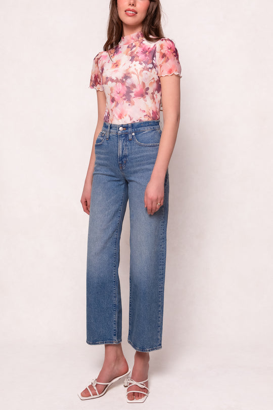 A woman stands against a light background wearing the Ivy City Co Ava Top in Purple Floral, high-waisted blue jeans, and white strappy sandals—a relaxed, maternity-friendly look for effortless style.