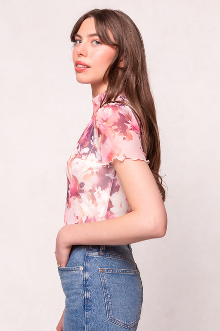 A woman with long brown hair stands in profile against a light background, wearing Ivy City Cos maternity-friendly Ava Top in Purple Floral and blue high-waisted jeans.