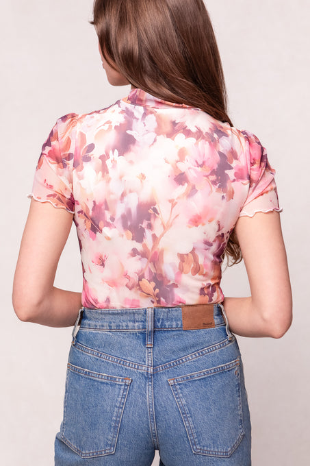 Woman with long brown hair, shown from the back, wears the Ivy City Co Ava Top in Purple Floral—a pink and purple sheer blouse—paired with high-waisted blue jeans. This outfit is stylish and maternity friendly.