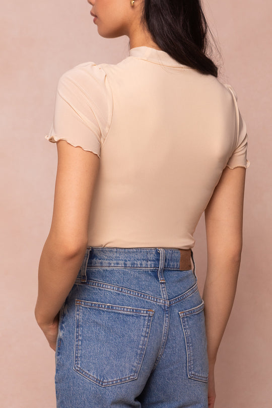 A woman stands with her back to the camera, wearing the Ava Top in Cream by Ivy City Co and high-waisted blue jeans against a soft beige background.