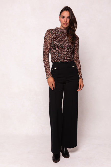 A woman stands against a light background, smiling softly. She wears the Ava Long Sleeve Top in Leopard by Ivy City Co, paired with high-waisted black wide-leg zippered pants and black ankle boots. Her long, wavy hair completes the look.