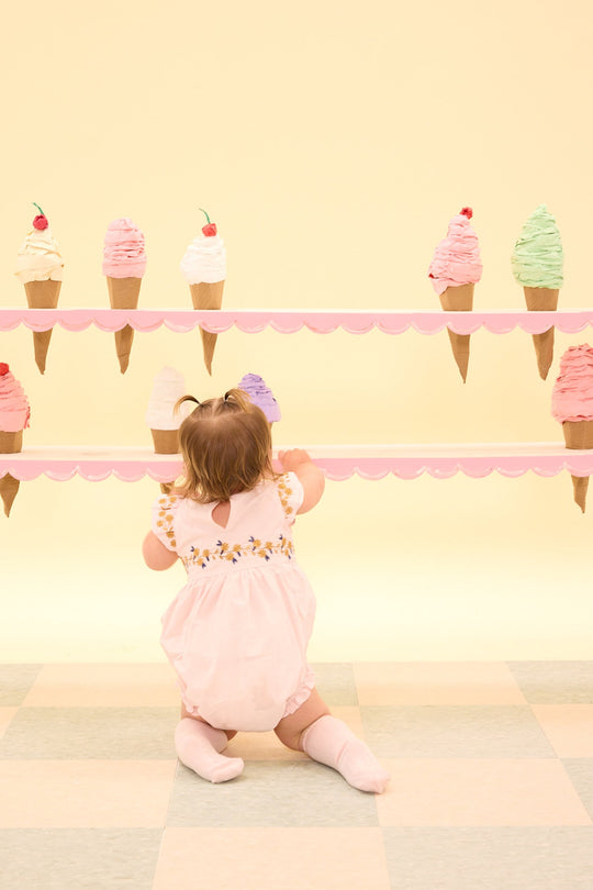 A young child in the Baby Hattie Romper in Embroidered Spring Floral by Ivy City Co sits on a pastel checkered floor, facing away and looking at two rows of colorful ice cream cones arranged against a yellow background.