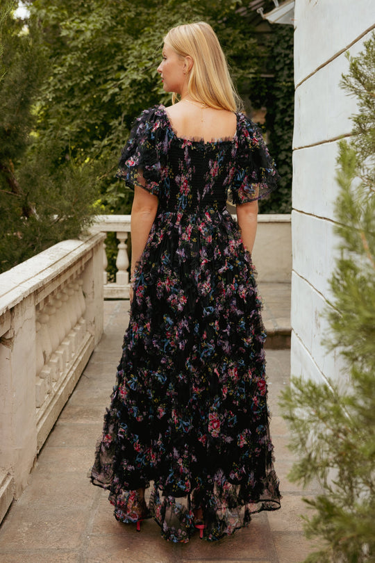A woman with blonde hair stands on a stone terrace, facing away. She wears the Florentina Dress in Obsidian Garden by Ivy City Co—a long black tulle floral gown with short puff sleeves and elegant ruffle details. Green trees and white balcony railings surround her.