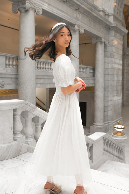 Wearing the Ivy City Co. Ballerina Dress in White and a headband, a woman stands on a marble staircase, her hair swaying as she turns. Behind her are grand stone columns and ornate architecture.