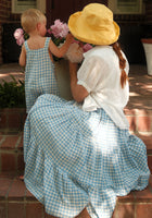 A woman wearing the Ivy City Co Zoey Skirt in Blue Gingham sits on steps with a child in matching overalls. They admire pink flowers together, sharing a sweet moment in their coordinated outfits.