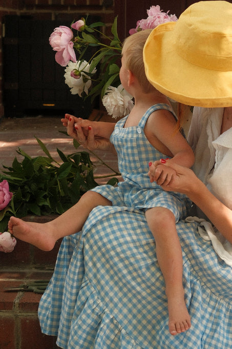 A woman in a yellow sunhat sits on brick steps, holding hands with a child wearing Ivy City Co's Baby Alex Overalls in Blue Gingham. Surrounded by pink and white flowers, the pair sits close, facing away from the camera.