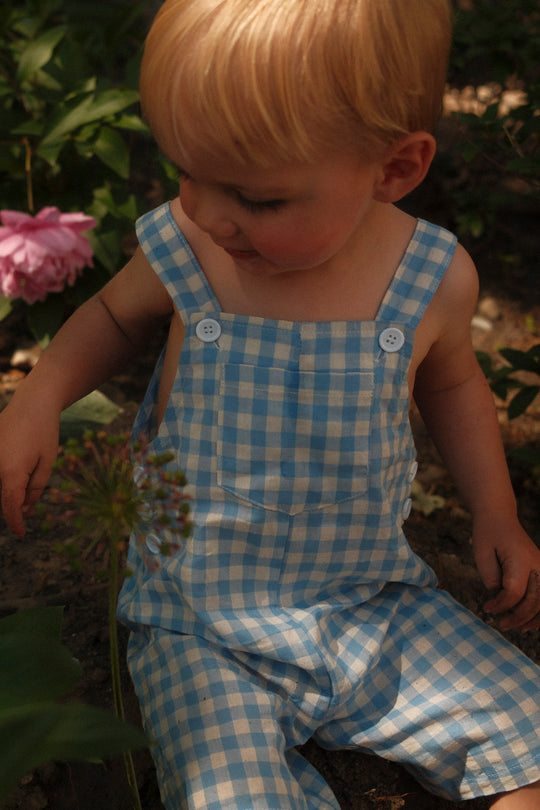 A young child wears Ivy City Co's Baby Alex Overalls in Blue Gingham, sitting outdoors among green plants and a pink flower, with sunlight softly illuminating their face.