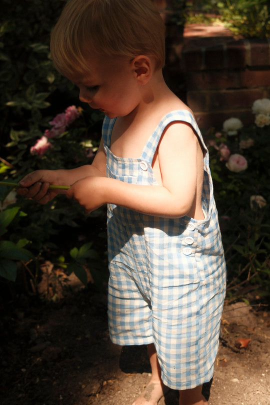 A young child with blonde hair stands barefoot in a sunny garden, wearing Ivy City Co’s Baby Alex Overalls in Blue Gingham—surrounded by flowers and greenery, holding a stick and gazing downward.