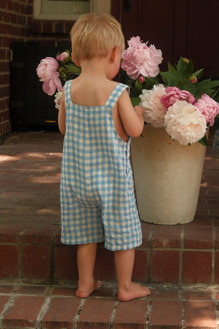 A young child wearing Ivy City Co’s Baby Alex Overalls in Blue Gingham stands barefoot on brick steps, leaning toward a large white pot of pink and white peonies—a perfect spring baby outfit moment.