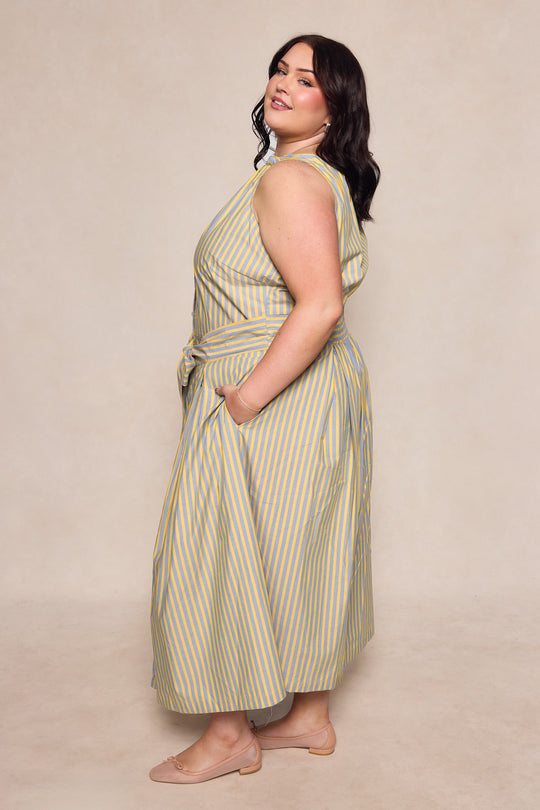 A woman poses in the Glen Dress in Green Stripe by Ivy City Co—a sleeveless, cotton ankle-length dress featuring green stripes, a tie at the waist, and convenient side pockets. She is standing sideways and wearing nude flats against a neutral background.