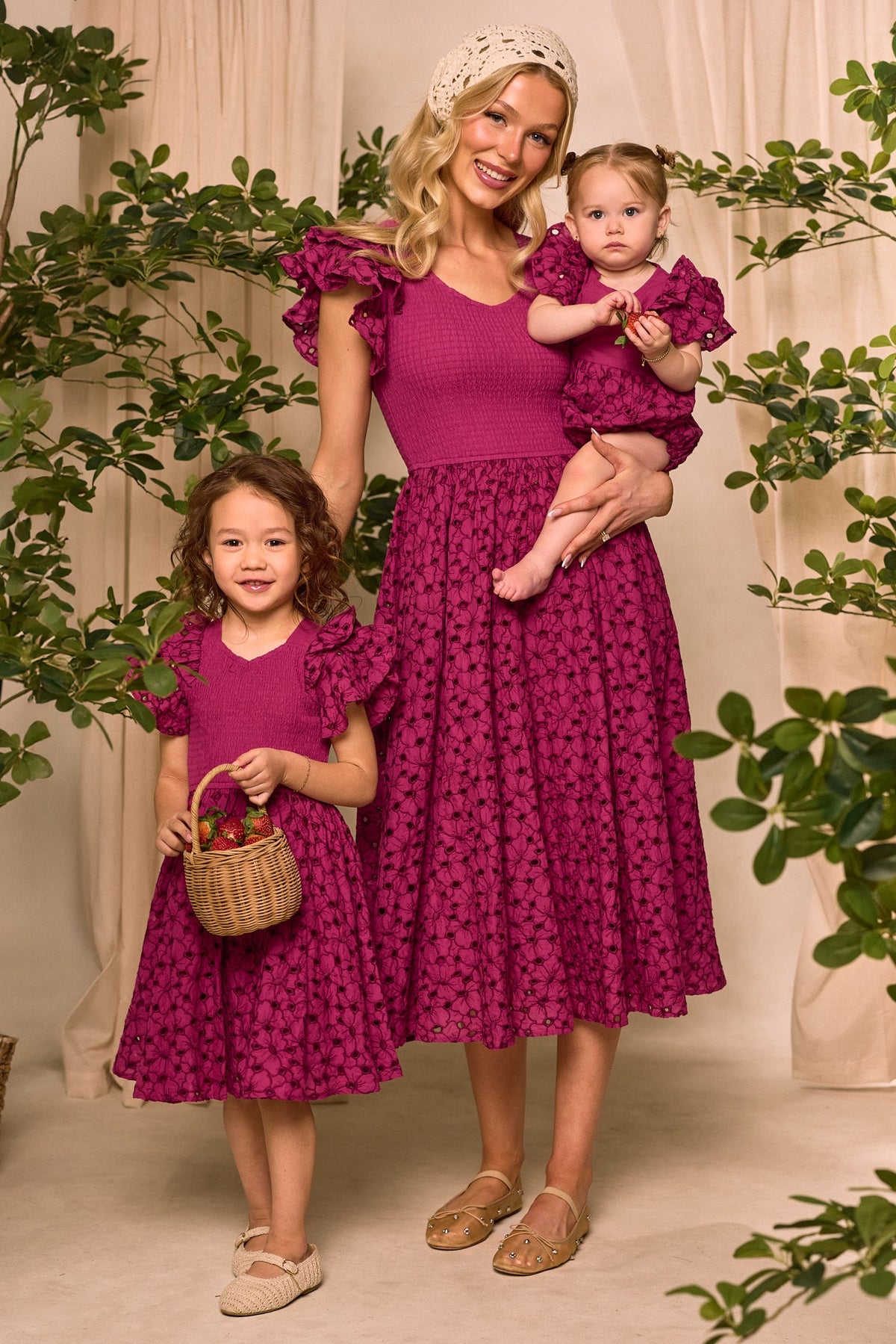 A woman and two young girls stand together, all wearing coordinating magenta dresses with floral patterns—an ideal choice for an Easter outfit. The woman holds the baby, who is dressed in the Ivy City Co Baby Hattie Romper in Magenta; the older girl carries a small basket. Green plants and beige curtains complete the background décor.
