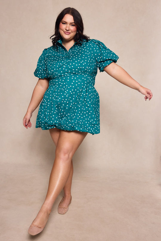 A woman poses confidently in the Ivy City Co Cody Dress in Deep Teal Polka Dot, a short dress featuring white polka dots and puff sleeves. She smiles with one foot forward, wearing nude flats against a neutral background.