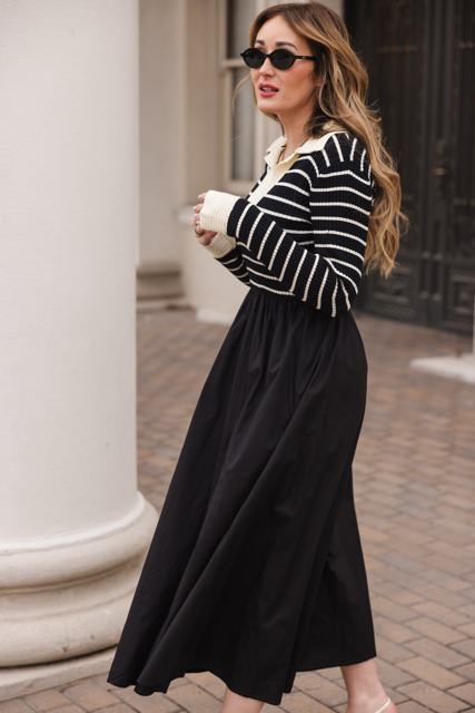 A woman in black sunglasses walks outdoors on a brick path near white columns, wearing the Ivy City Co Adelina Dress in Black—a chic black and white striped knit bodice long-sleeve dress with a long black skirt.