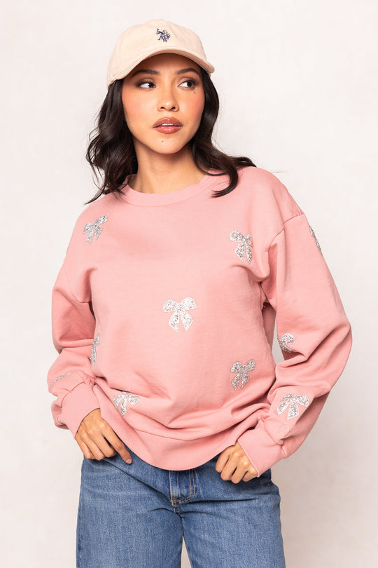A woman wearing the Micah Crew Neck Sweatshirt in Pink & Bows by Ivy City Co, styled with a coordinating light pink cap and blue jeans, stands against a neutral background with one hand in her pocket.