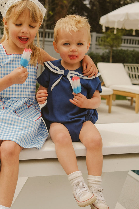 Two young children sit by a pool, smiling and holding red, white, and blue popsicles. One wears a blue gingham dress, while the other looks adorable in the Baby Sailor Romper in Navy from Ivy City Co. Lounge chairs and an umbrella are in the background.
