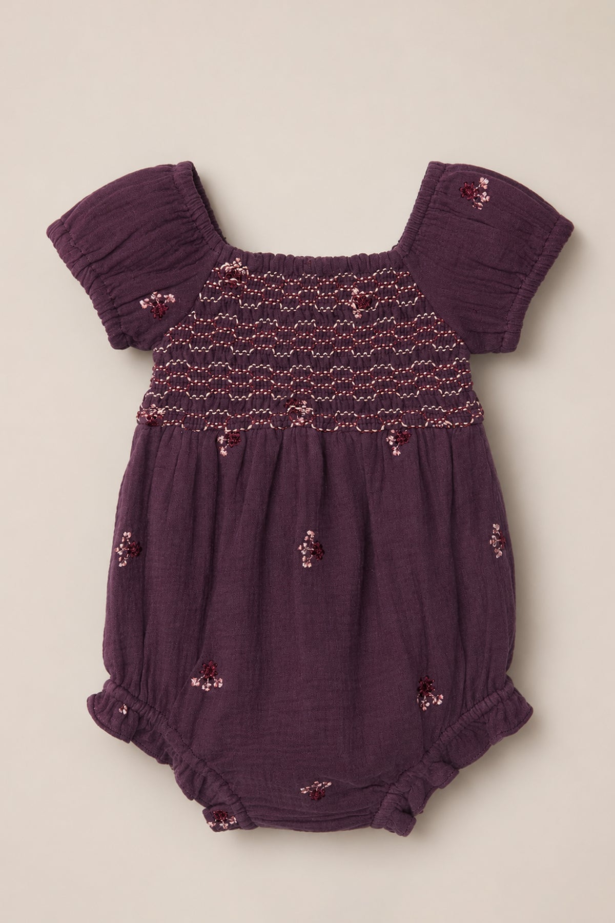 The Ivy City Co Baby Acantha Romper in Aubergine Embroidered Gauze showcases charming puff sleeves, a beautifully smocked bodice, elastic leg openings, and delicate floral embroidery scattered across the fabric. It is laid flat on a neutral background for a sweet and timeless presentation.