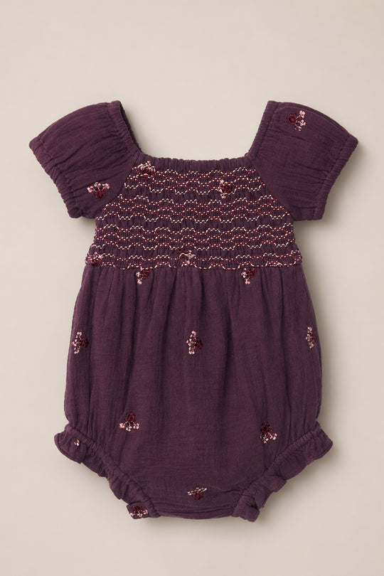 The Ivy City Co Baby Acantha Romper in Aubergine Embroidered Gauze showcases charming puff sleeves, a beautifully smocked bodice, elastic leg openings, and delicate floral embroidery scattered across the fabric. It is laid flat on a neutral background for a sweet and timeless presentation.