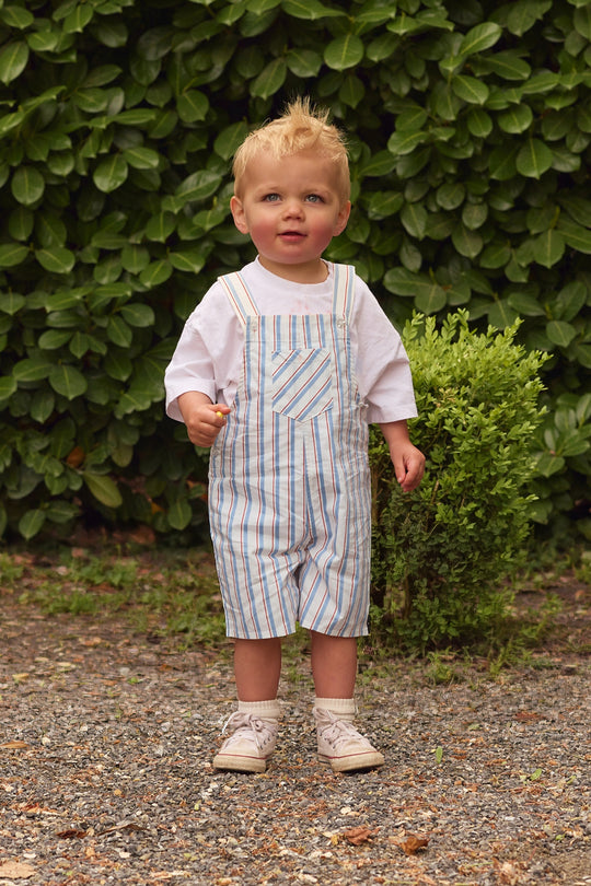 A young toddler with short blond hair stands on gravel by green bushes and a small shrub, wearing a white shirt, beige shoes, and Ivy City Co's Baby Alex Overalls in Heritage Stripe.
