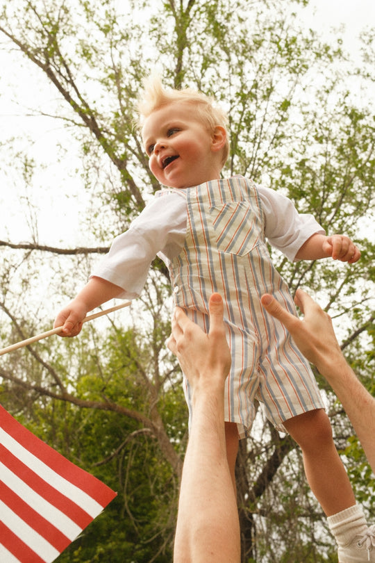 A smiling toddler in Ivy City Co's Baby Alex Overalls in Heritage Stripe is lifted outdoors by adult hands, holding a small American flag—an adorable Fourth of July look with green trees and a cloudy sky behind.