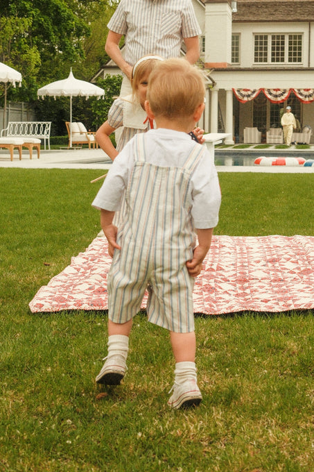 A toddler in Ivy City Co’s Baby Alex Overalls in Heritage Stripe stands on grass near a patterned blanket, facing two adults and a child by a pool with patriotic bunting—an ideal outfit for the Fourth of July.
