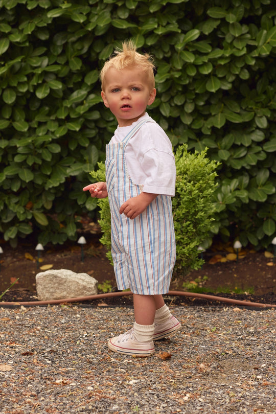 A young child with blond hair stands outdoors on gravel, wearing a white t-shirt and Ivy City Co Baby Alex Overalls in Heritage Stripe, paired with socks and sneakers. A lush green hedge and a small bush are in the background.