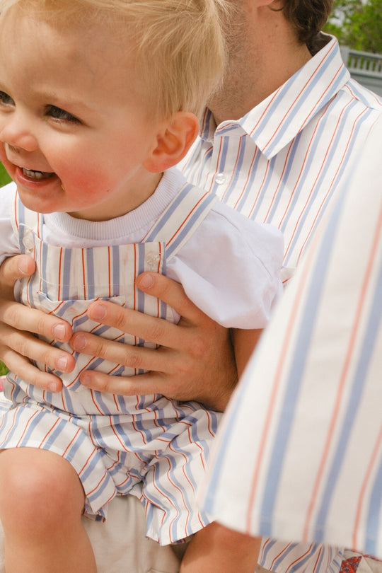 A smiling child wears Ivy City Co's Baby Alex Overalls in Heritage Stripe, held by an adult in a matching shirt. The child's rosy cheeks and only part of the adult’s face are visible; another person in similar festive attire is partly seen.