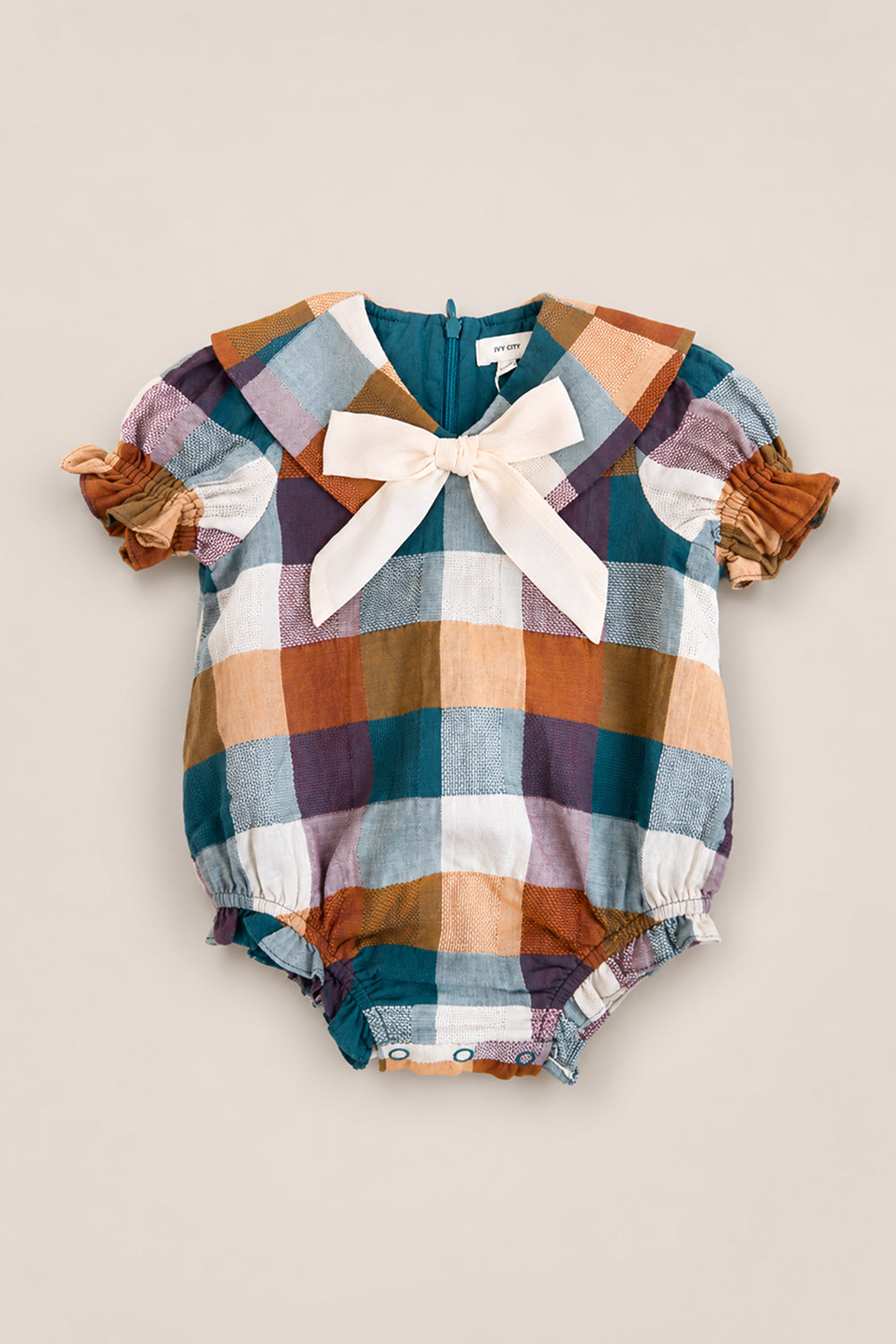 The Baby Alice Romper in Maize Gingham by Ivy City Co is a charming plaid cotton-blend romper featuring puff sleeves, a large sailor-style collar, a cream bow at the neckline, and gathered leg openings. The fabric showcases teal, brown, purple, and white checks for a classic touch.