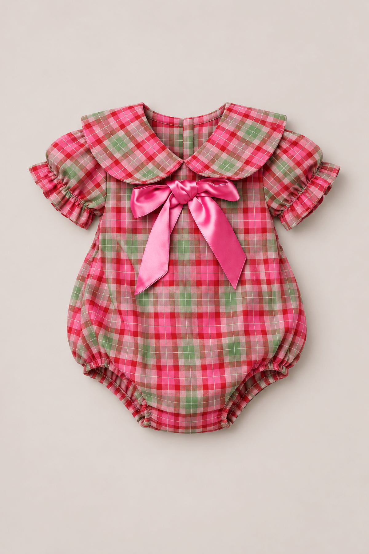 The Baby Alice Romper in Rosewood Tartan by Ivy City Co features pink, green, and white cotton plaid with puff sleeves, a Peter Pan collar, and a large shiny pink bow on the chest. Designed for both comfort and style, this charming romper includes elastic around the legs and convenient back buttons.