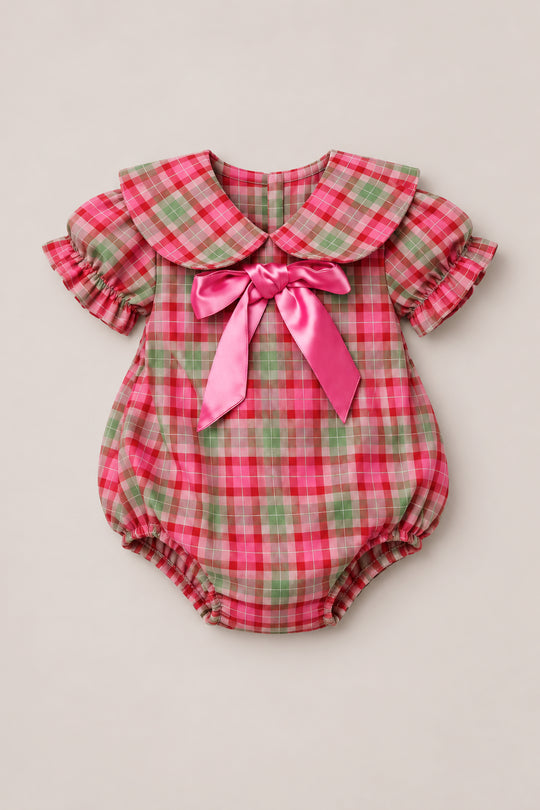 The Baby Alice Romper in Rosewood Tartan by Ivy City Co features pink, green, and white cotton plaid with puff sleeves, a Peter Pan collar, and a large shiny pink bow on the chest. Designed for both comfort and style, this charming romper includes elastic around the legs and convenient back buttons.