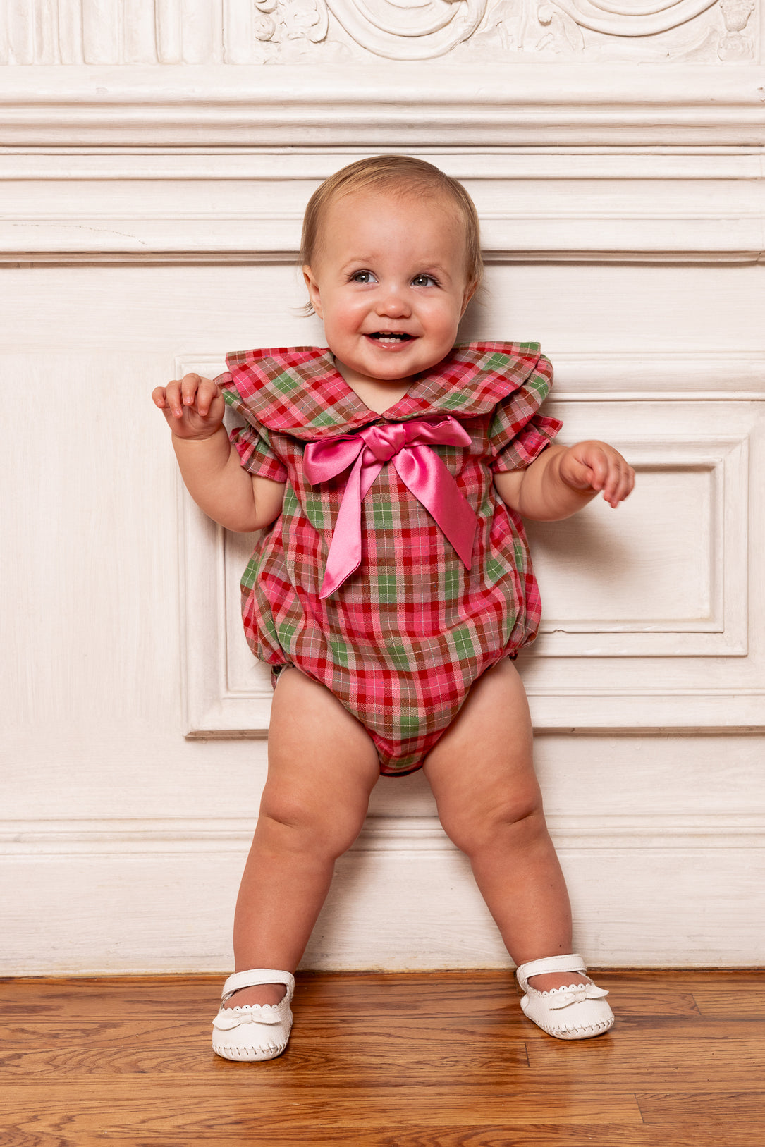 Baby wearing a red plaid outfit with a pink bow standing against a decorative wall.