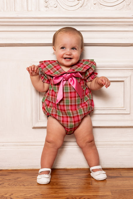 Baby wearing a red plaid outfit with a pink bow standing against a decorative wall.