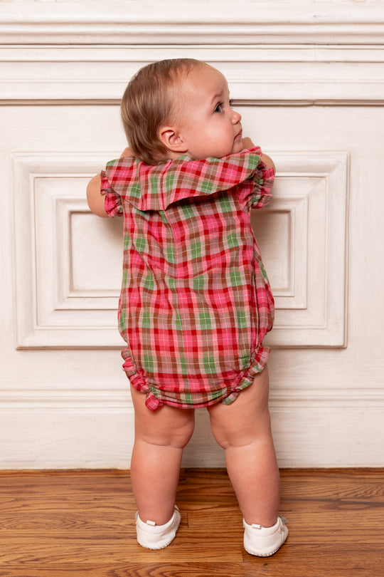 Baby in a red and green checkered outfit standing against a white wall.