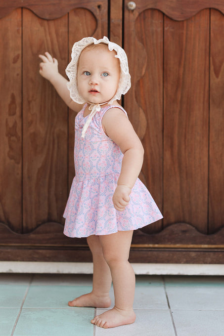 A baby wearing the Baby Bitsy Swim Dress in Raquel Pink Floral and a white bonnet stands barefoot on a tiled floor, facing a wooden cabinet with one hand touching it and looking toward the camera.