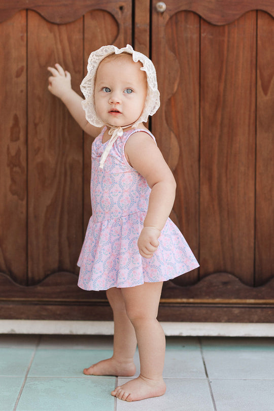 A baby wearing the Baby Bitsy Swim Dress in Raquel Pink Floral and a white bonnet stands barefoot on a tiled floor, facing a wooden cabinet with one hand touching it and looking toward the camera.