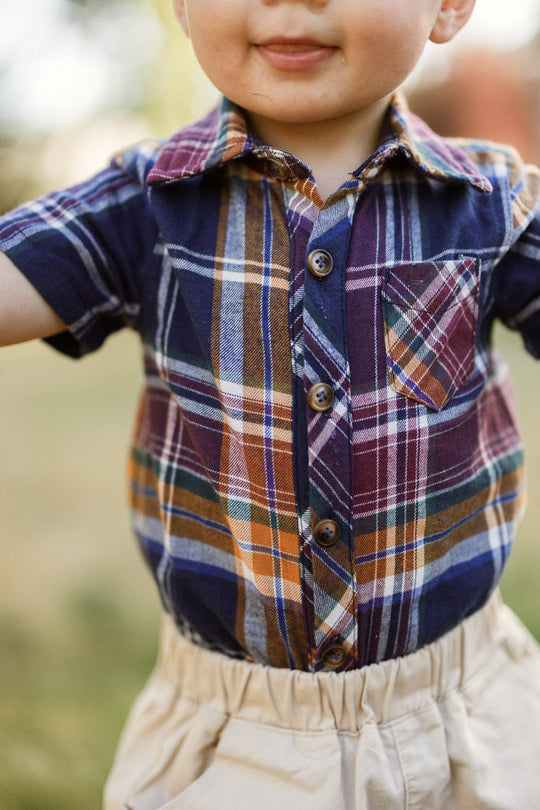 A young child stands outdoors, smiling with arms slightly outstretched, wearing the Ivy City Co Baby Boy James Romper in Midnight Plaid.